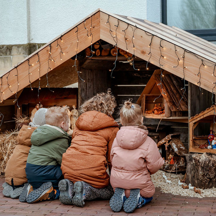 4 kinderen knielen voor kleine kerststalletjes die in een grotere kerststal zijn geplaatst