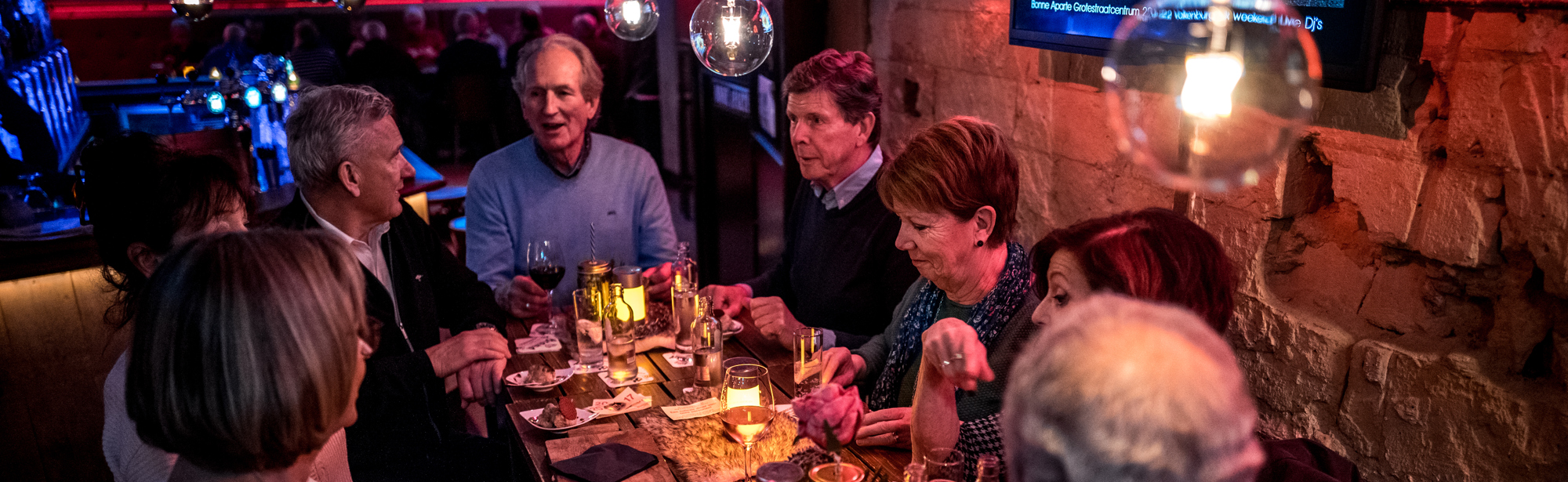 8 mensen zitten aan een lange houten tafel met hapjes en drankjes