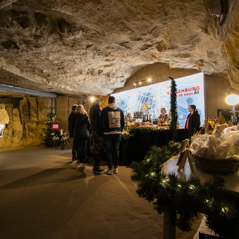 Mensen staan bij een kertstkraampje in een mergelgrot met daarachter een grote stadsplattegrond van valkenburg