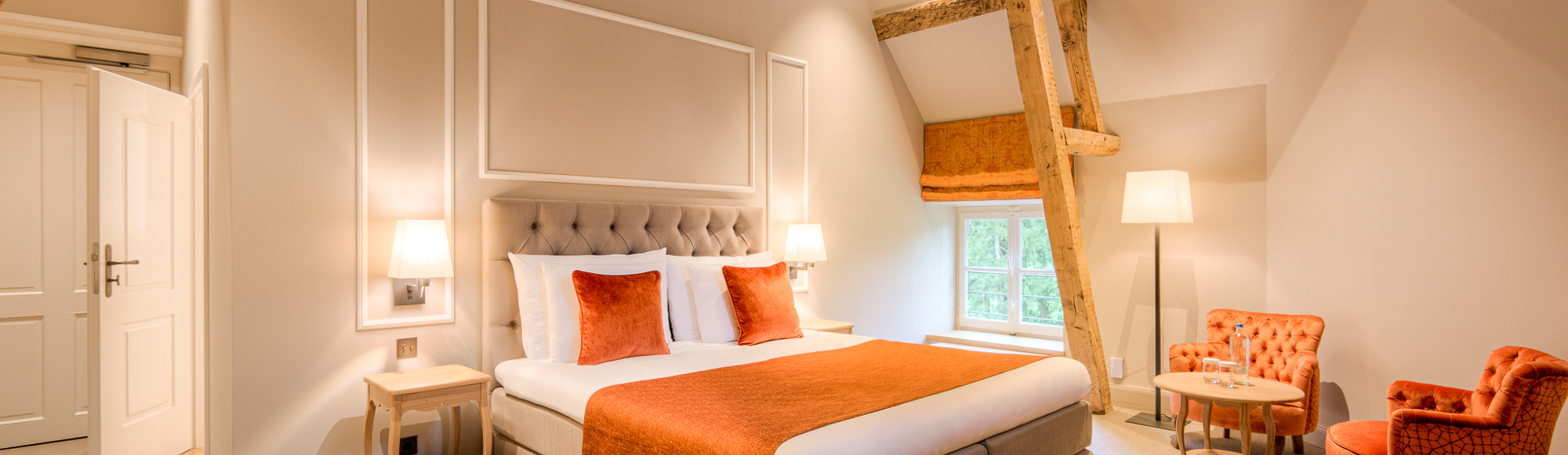 Een hotelkamer met oranje details op het bed en twee oranje stoeltjes. 